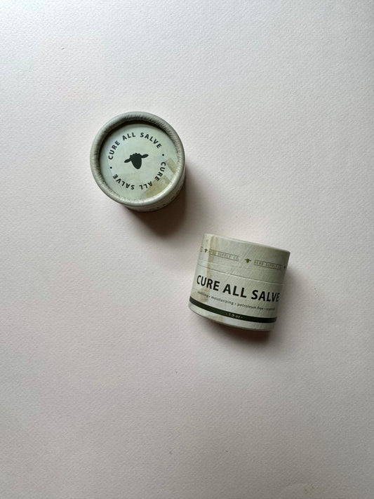 Two small Cure All Salve jars on a plain off-white background, with one jar showing its lid and the other showing the labeled body.